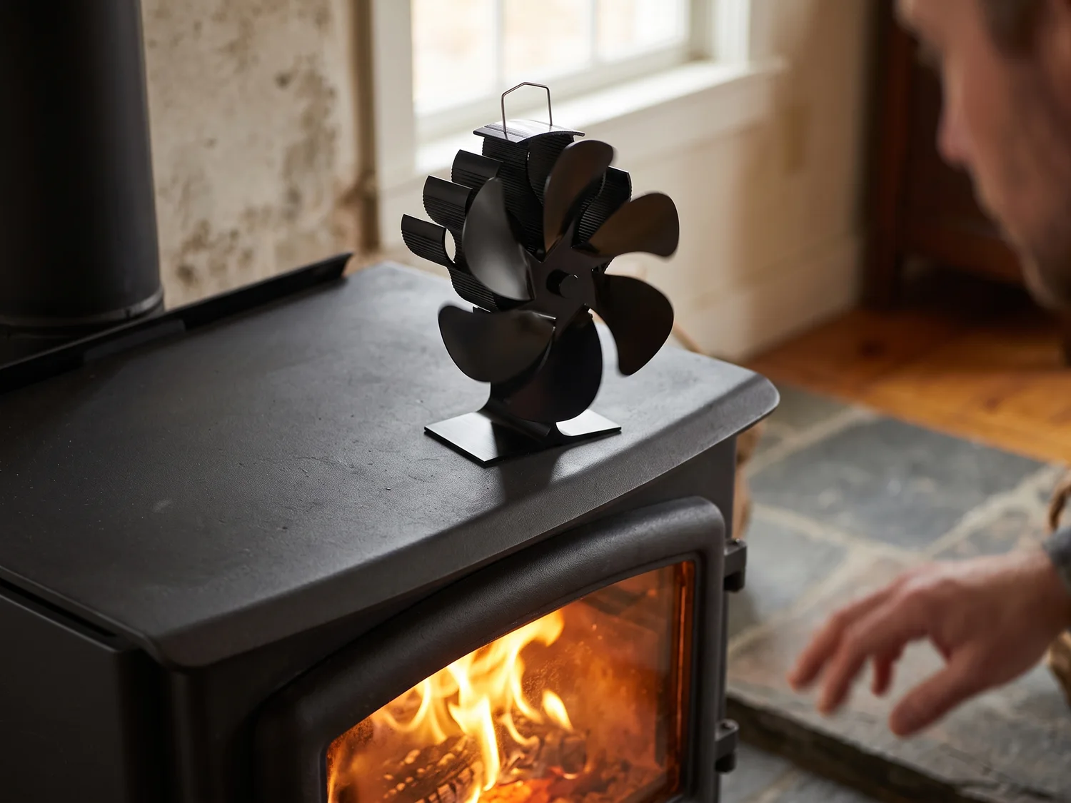 A small black six-vane fan turning on the flat top of a cast iron wood stove, firelight in the glass door below